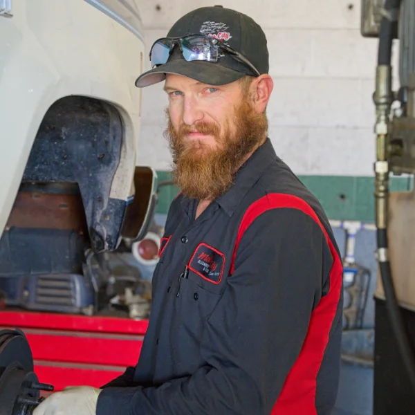 A mechanic with a red beard wearing a black and red uniform and gloves works on a vehicle in an auto repair shop.